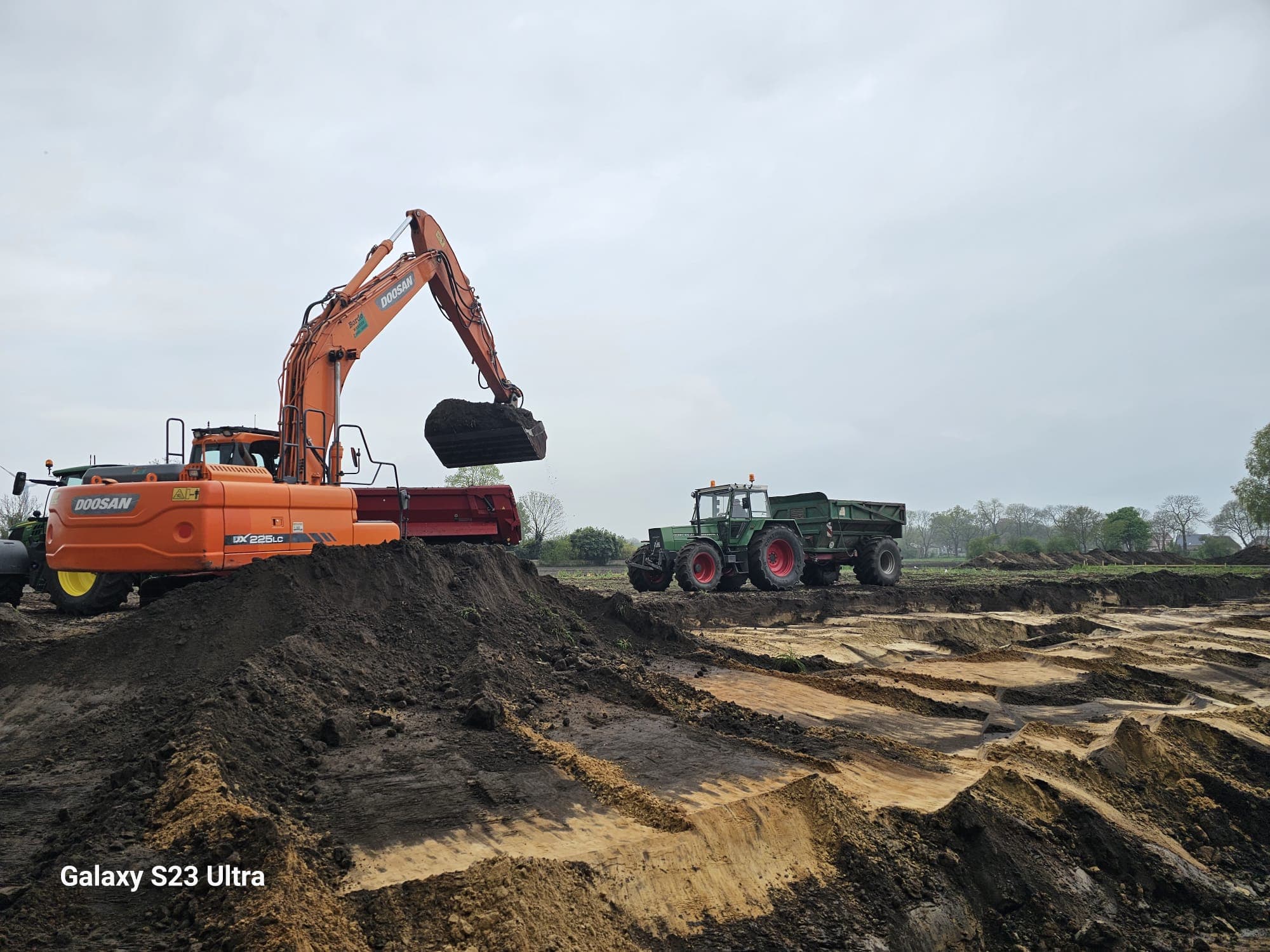 Doosan-Bagger belädt einen Stalldungstreuer, im Hintergrund ein Traktor mit Anhänger auf dem Feld.