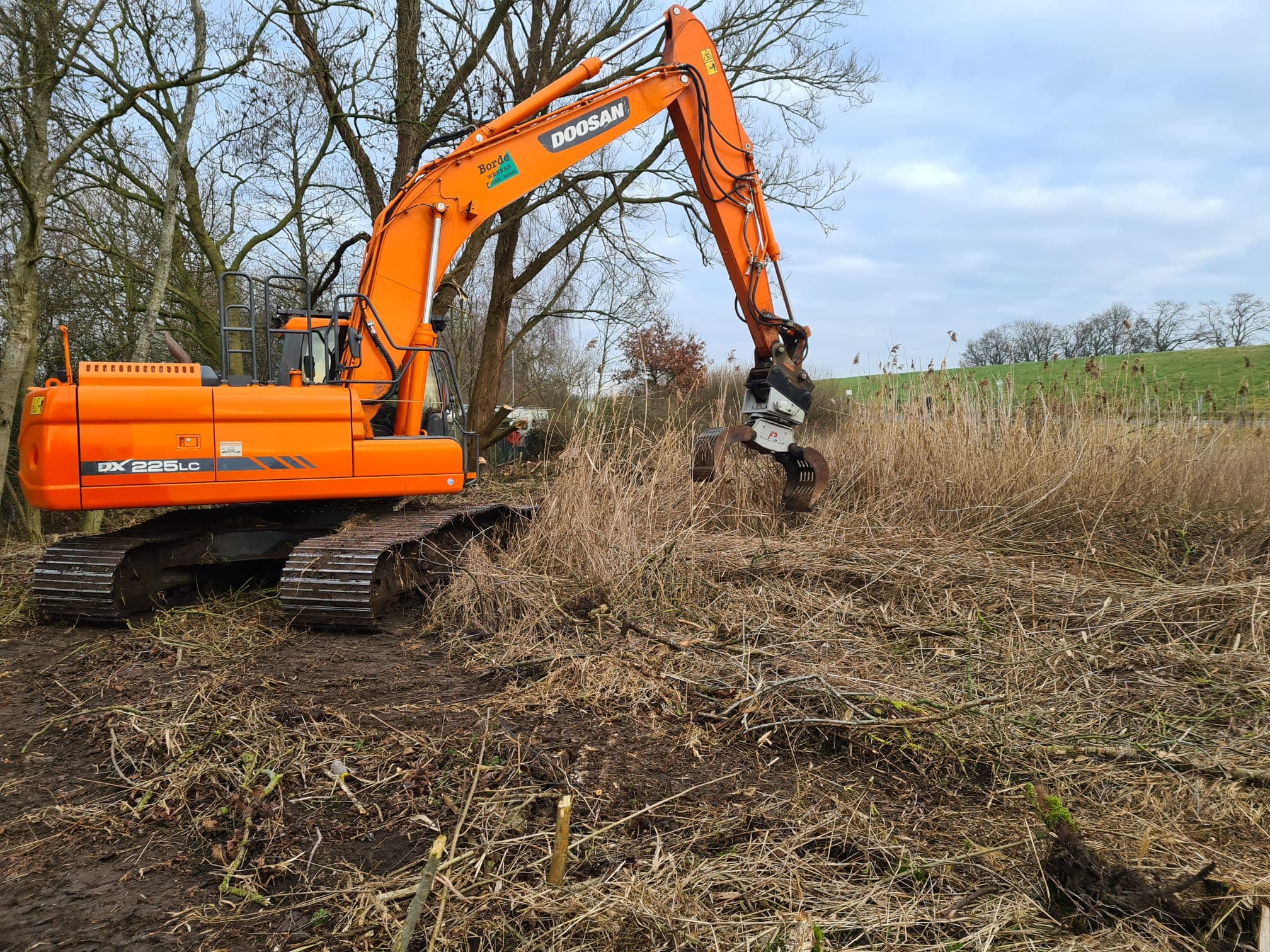 Bagger mit Grapple räumt Gehölz und Reisig entlang eines Grabens.
