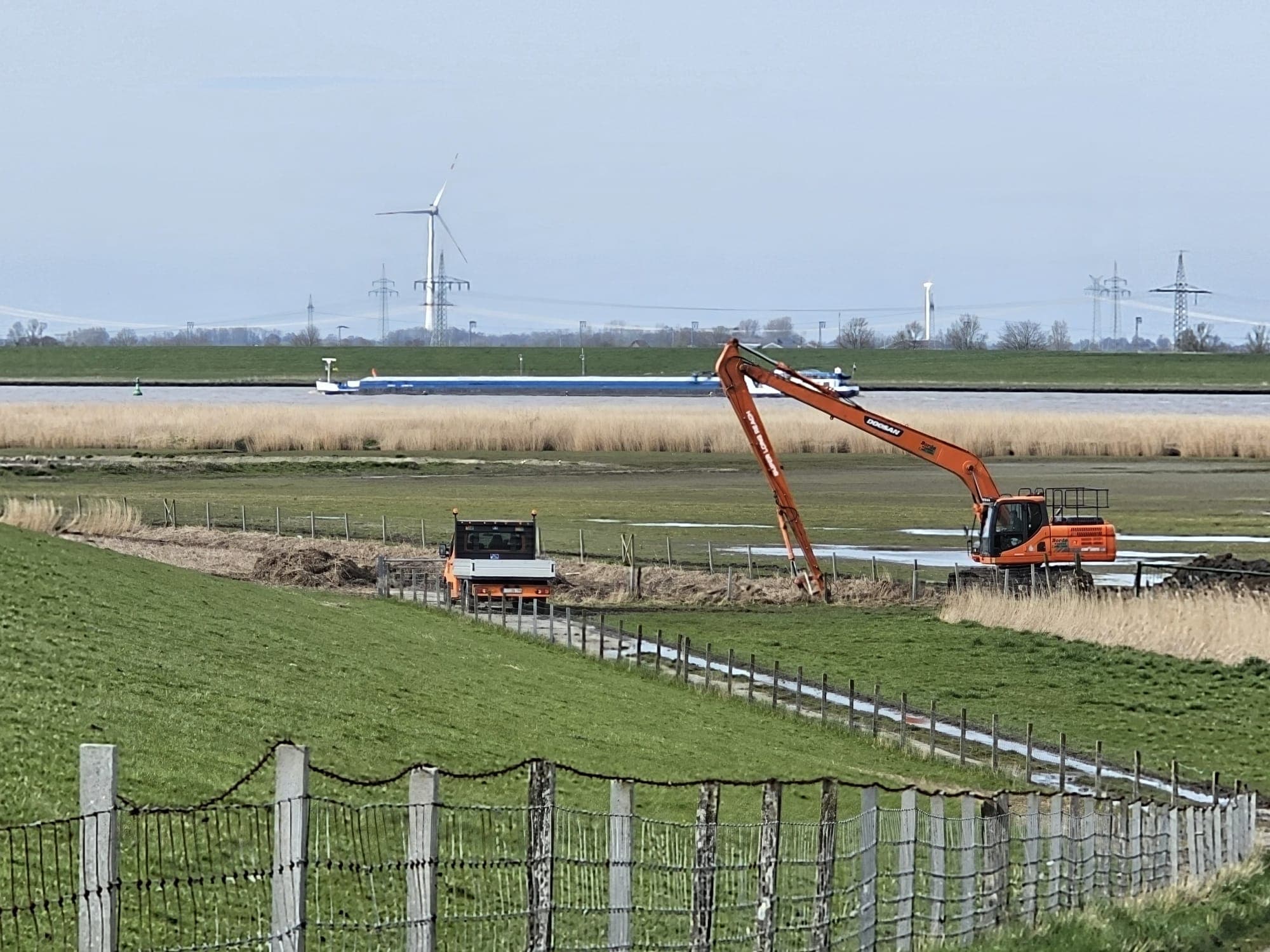 Langarmbagger von Landschaftpflege Borde bei der Grabenreinigung am Deich, im Hintergrund ein Frachtschiff und ein Windrad.