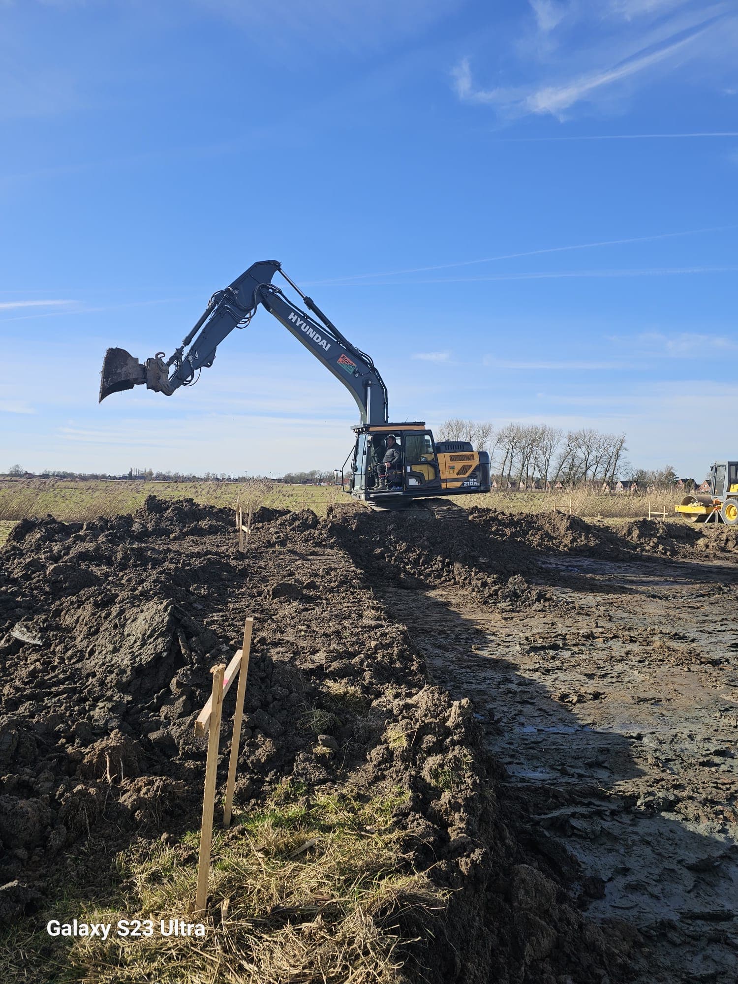 Hyundai-Bagger hebt Erdaushub bei Grabenarbeiten auf einer Baustellenfläche, im Hintergrund eine Walze.