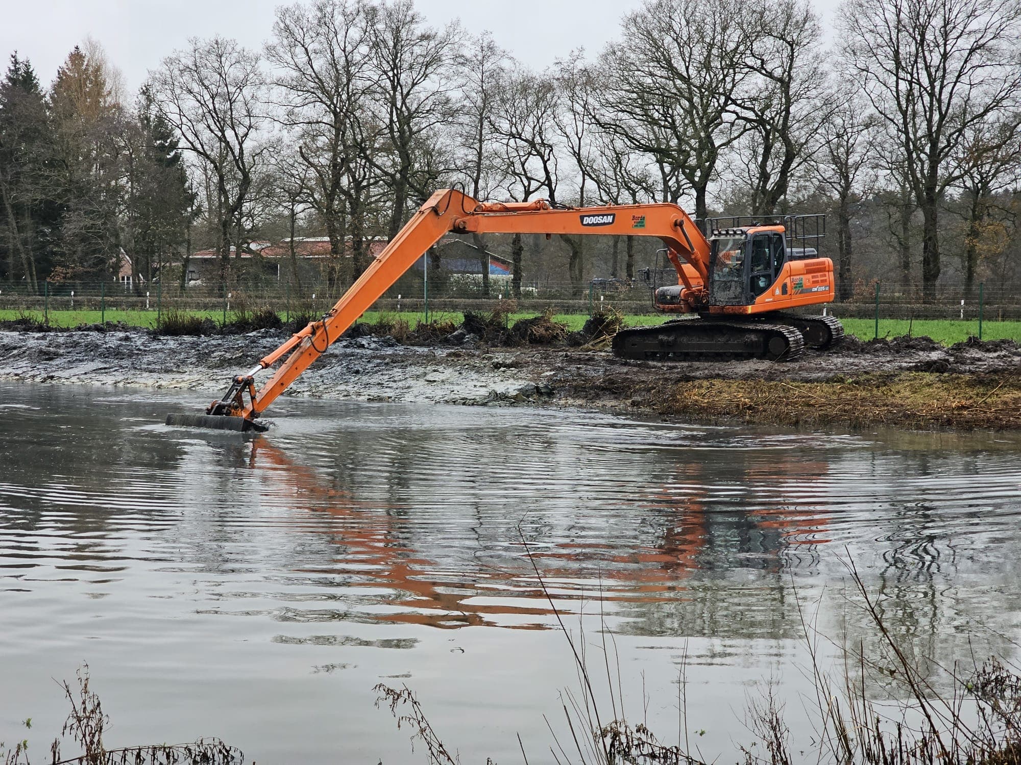 Langarmbagger reinigt einen wassergefüllten Graben, die Baggerschaufel arbeitet direkt in der Wasserfläche.