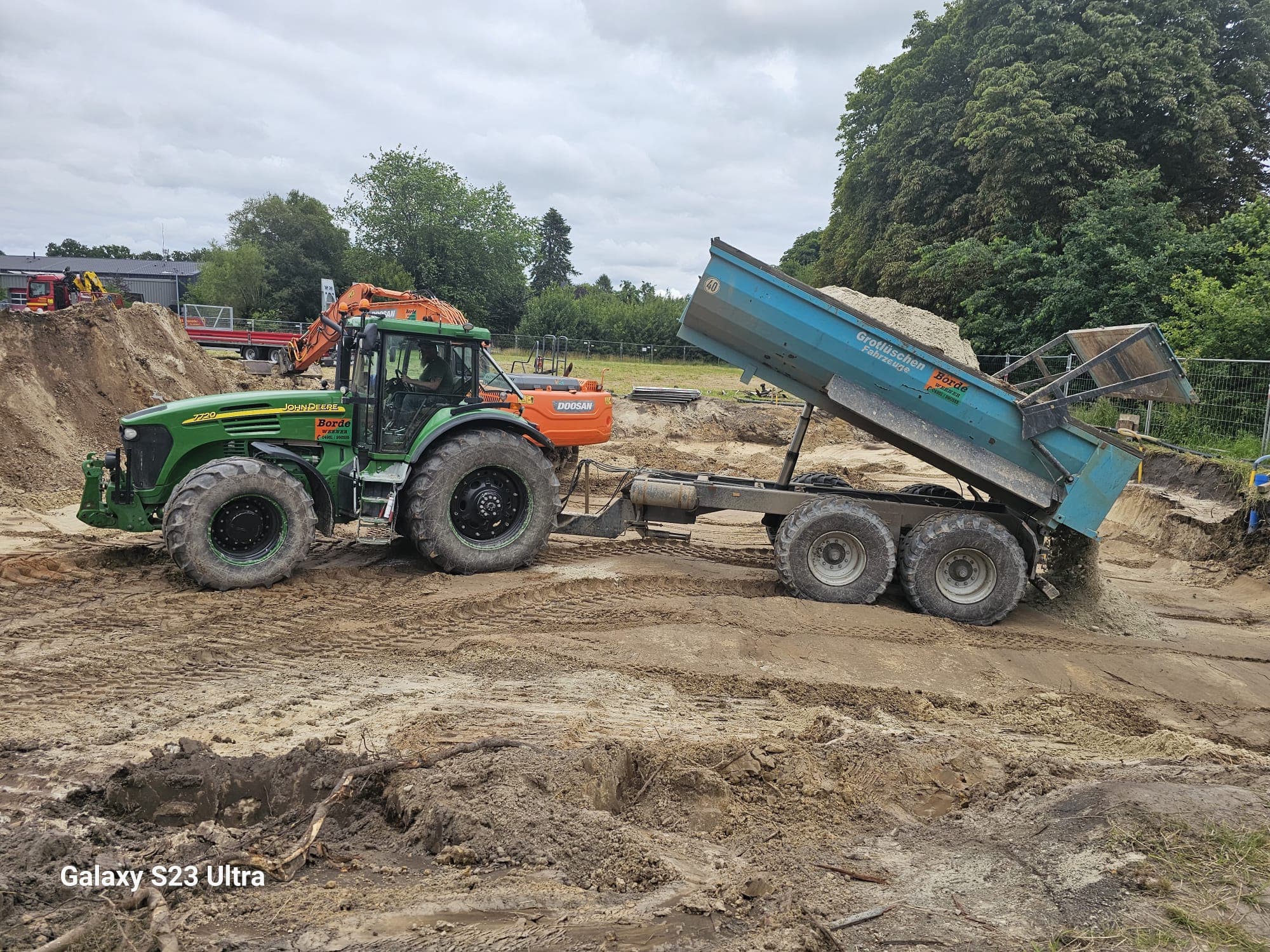 John-Deere-Traktor mit Kippanhänger (Borde Weener-Logo) kippt Sand auf einer Baustelle ab, im Hintergrund ein Bagger.